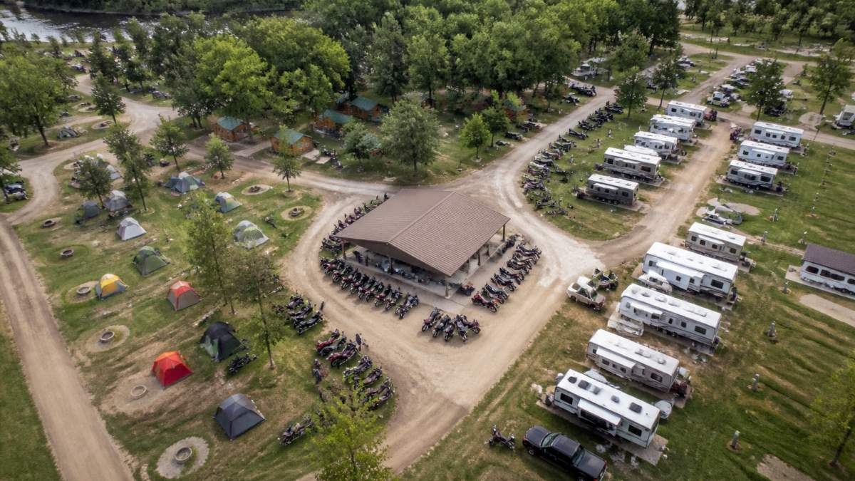 Aerial view of River's Edge Campground showing organized tent camping rows with fire pits, RV hookup stations with utility pedestals, cabin structures nestled among trees, and a central gathering pavilion with motorcycle parking areas clearly marked