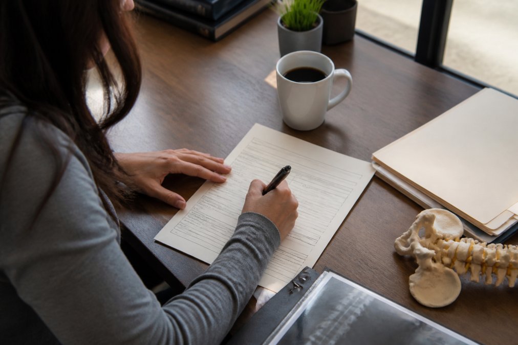 A patient sitting at a desk completing a health history form with a pen, with medical documents and a cup of coffee nearby