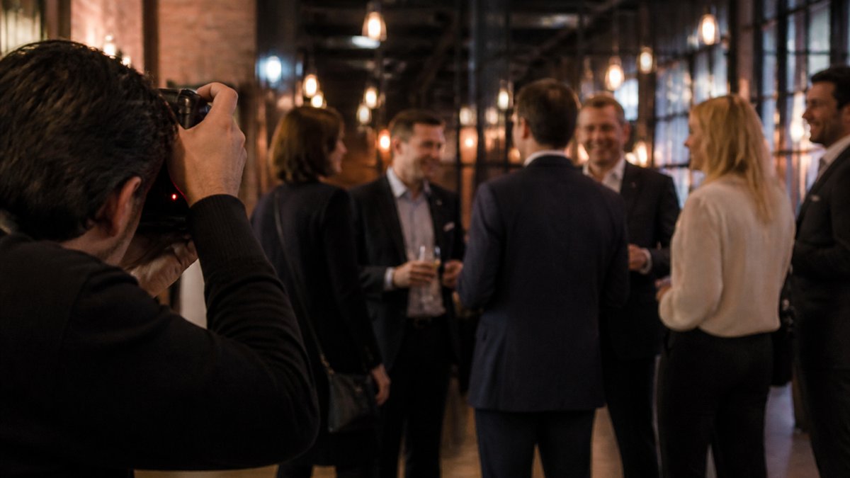Professional photographer discreetly capturing candid moments as a group of corporate professionals engage in animated conversation at a reception, with warm ambient lighting and natural interaction in the background