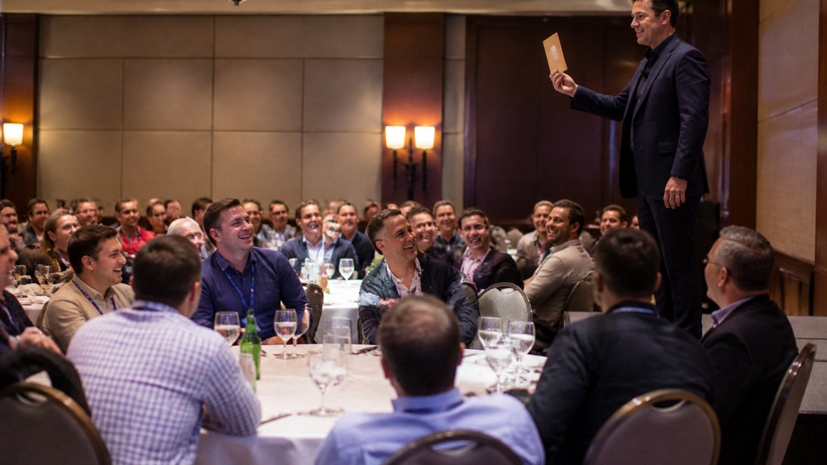 A corporate audience in a hotel ballroom laughing and leaning forward as a mentalist on stage holds up a sealed envelope, with guests seated at round tables in the foreground