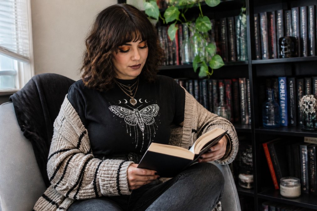 Young adult styled in layered bookish apparel including a graphic tee, oversized cardigan, and custom accessories, posed confidently in natural window lighting with books visible in the background