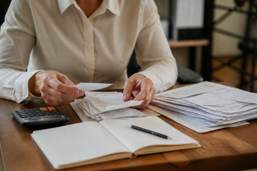 Close-up of hands organizing financial documents and receipts on a desk with a calculator and open notebook, warm office lighting suggesting careful and methodical record-keeping