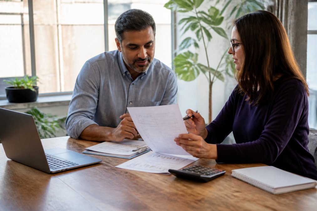 Small business owner and professional bookkeeper reviewing financial documents together at a wooden desk in a bright office, laptop open with accounting software visible