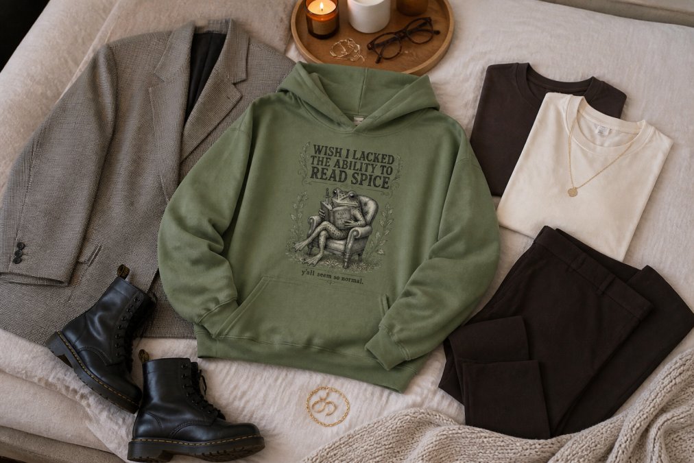 Flat lay of coordinated booktok-inspired clothing pieces arranged on a neutral surface, including layered graphic tees, oversized hoodie, structured blazer, tailored trousers, and accessories like chains and rings, with soft natural lighting from above