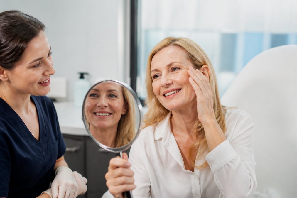 A professional woman in her late 40s looking into a mirror in a bright, modern medical office, smiling naturally, with a medical professional standing nearby