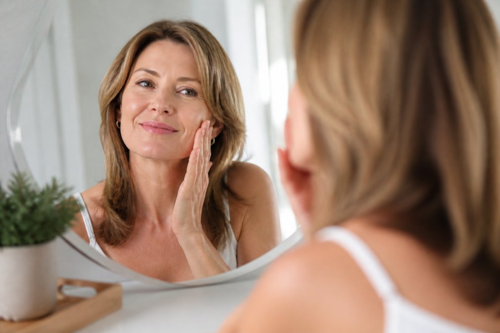 A woman in her 40s looking into a mirror, gently touching her cheek and smiling, with a clean, bright clinic background and soft natural lighting
