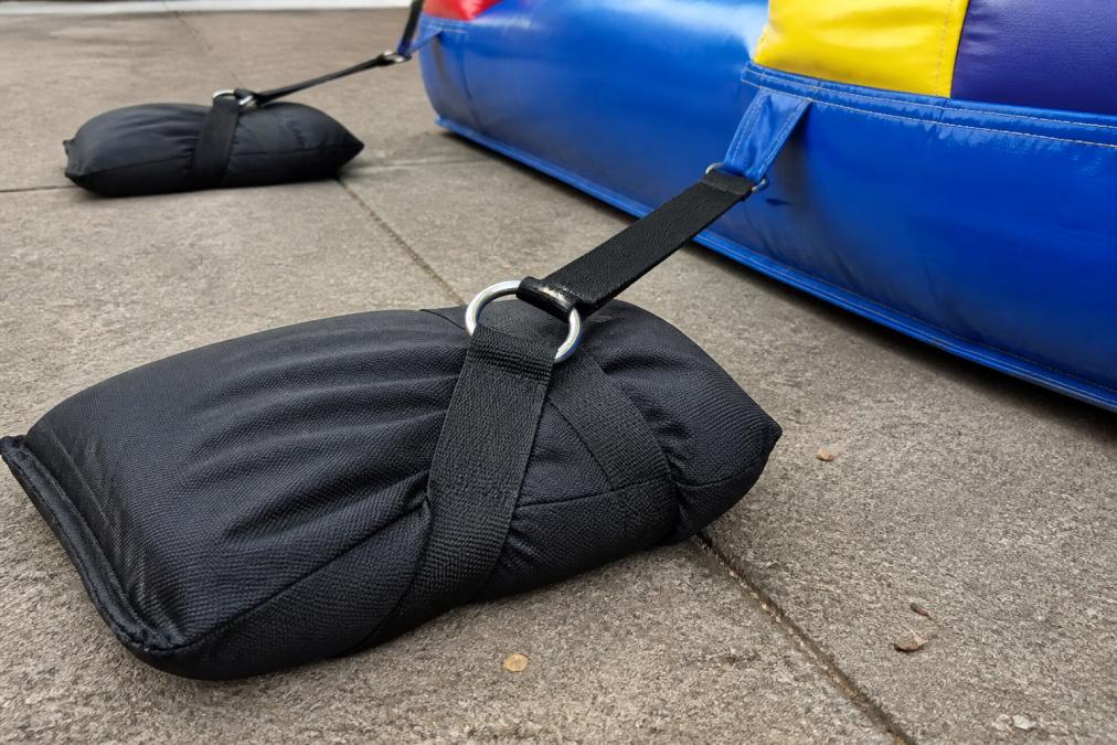 Close-up of heavy-duty sandbag anchors with D-ring attachment secured to bounce house tie-down straps on concrete surface, showing proper weight distribution and secure fastening