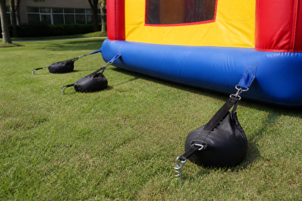 Close-up of sandbags and ground stakes properly securing an inflatable bounce house to level grass, showing multiple tethering points and clearance around the structure