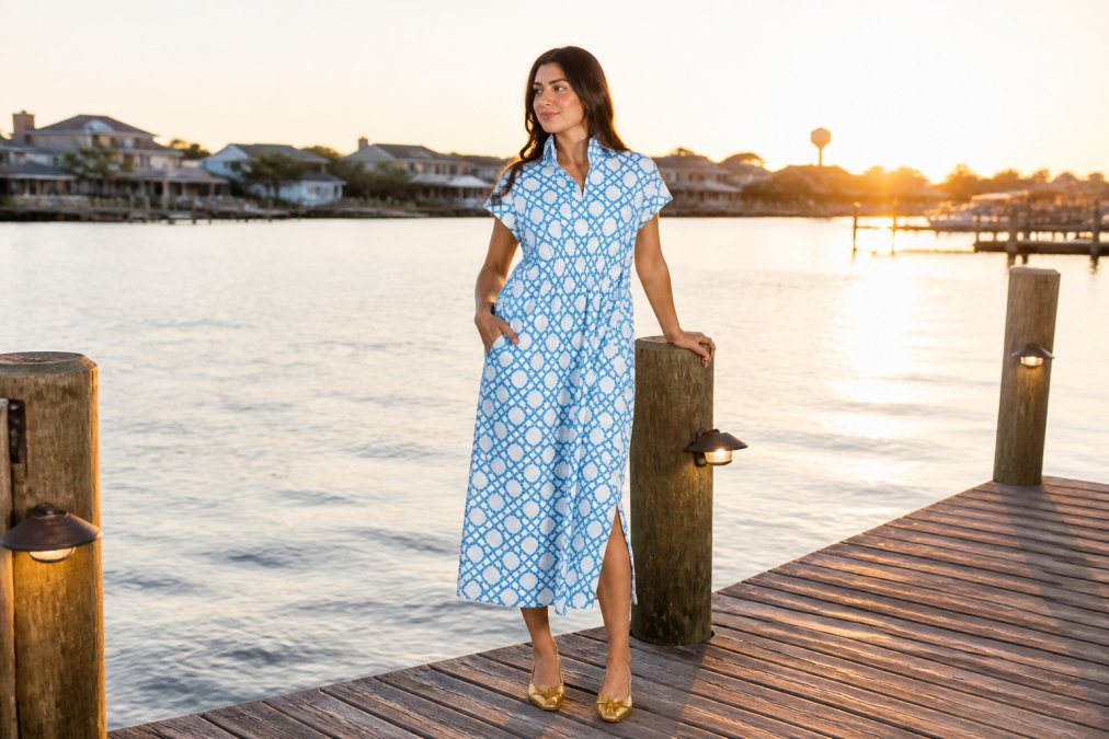 Woman wearing a flowing coastal midi dress in soft blue tones standing on a wooden deck overlooking calm water at golden hour, embodying effortless elegance and seaside-inspired style