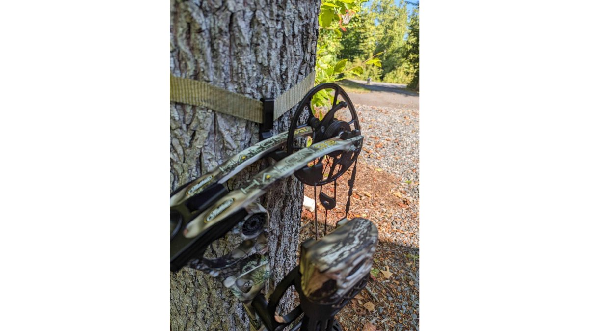 A saddle hunter hanging a compound bow on a modular bow hanger attached to a tree, with a gear strap and hunting pack visible, early morning light in a hardwood forest