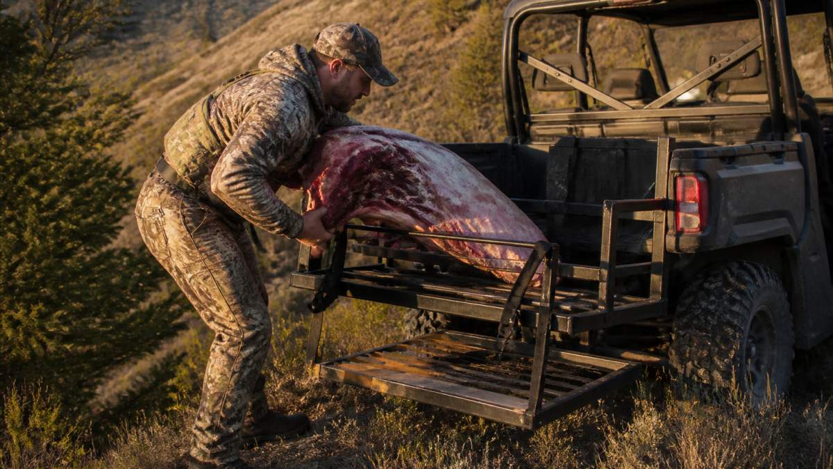 A solo hunter in camo loading an elk quarter onto a UTV-mounted game hauler on a steep, brushy hillside in late afternoon light
