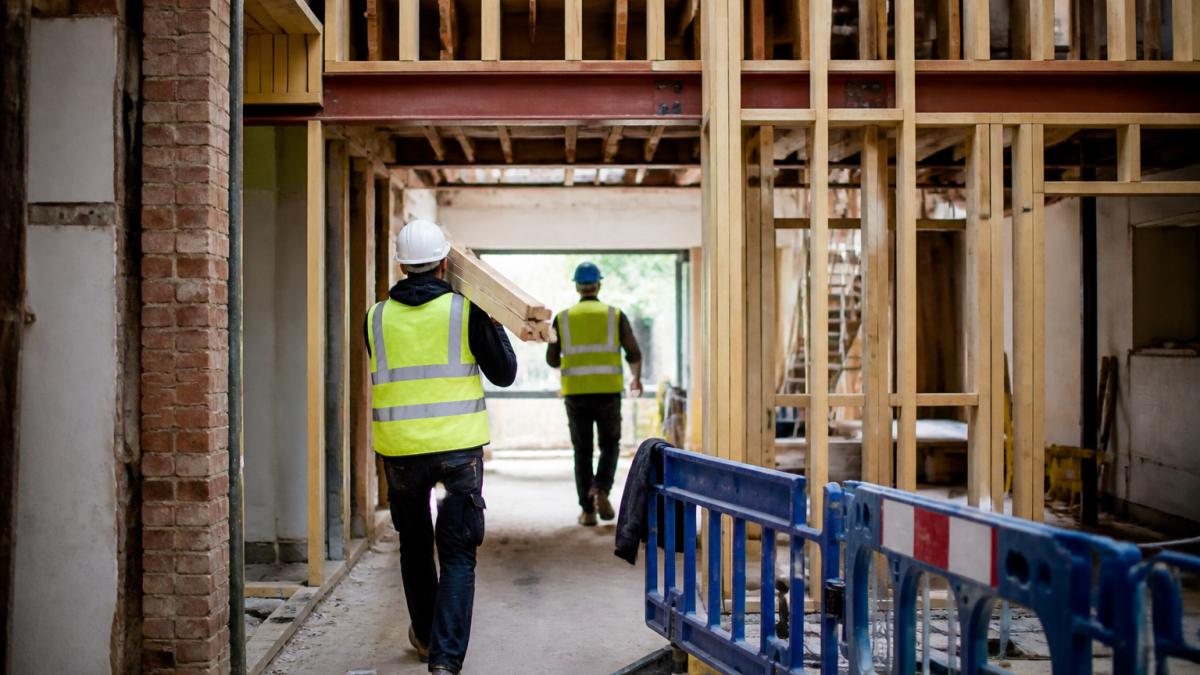 Active renovation site with workers carrying materials through protective barriers, partially completed structural framework visible with exposed beams and safety equipment positioned throughout