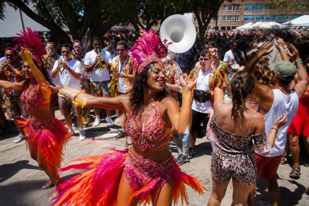 Brassband optredend op openluchtfestival met blazers, drummers en danseressen in volle energie, publiek dansend in zomerzon, felgekleurde kostuums en beweging