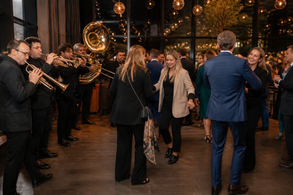Professionele brassband optredend op elegant bedrijfsfeest in moderne zalenruimte, muzikanten in zwart kostuum met blazers, gasten in zakelijke kleding dansend en lachend, warme verlichting