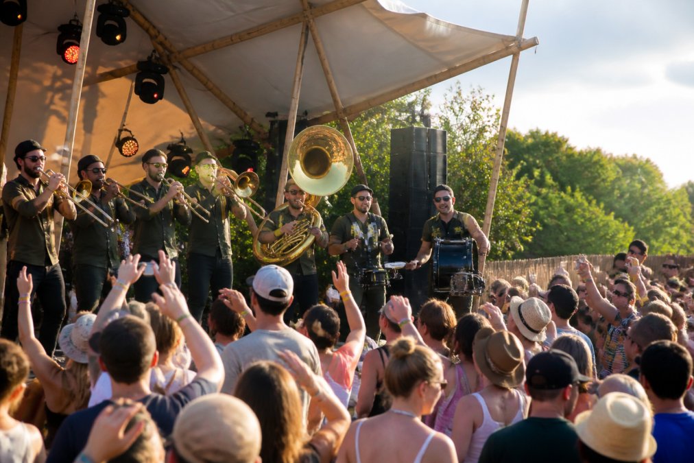 Energieke brassband met blazers en percussionisten die live optreden op een buitenpodium voor een groot, dansend publiek op een zomerfestival, met felle zonneschijn en enthousiaste bezoekers op de voorgrond