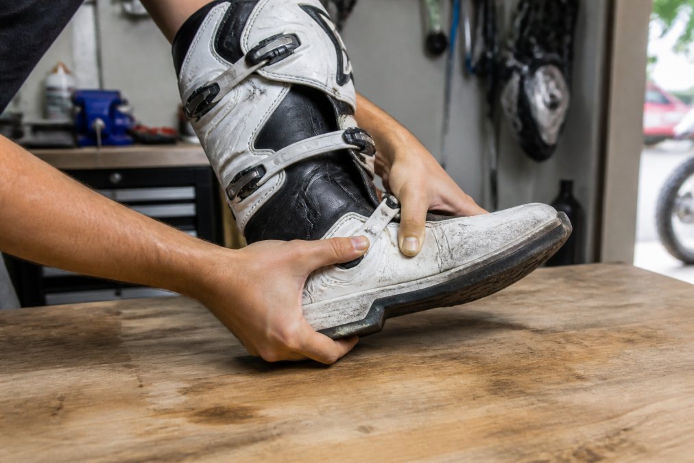 Hands demonstrating manual flexing technique on a motocross boot resting on a workbench, showing proper ankle flex zone pressure with natural workshop lighting