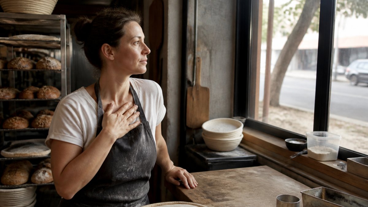 Professional at desk pausing mid-work, hand on chest, looking out the window with a calm, grounded expression in natural light