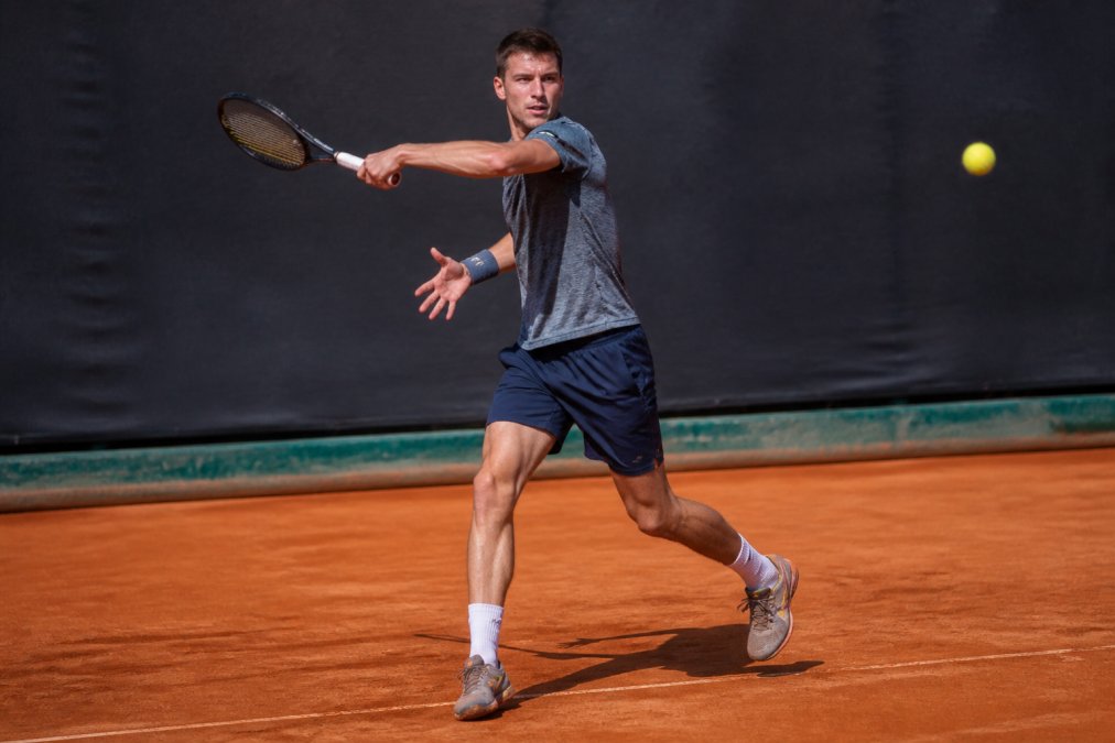 Competitive tennis player executing a forehand stroke on a clay court with proper footwork positioning and weight transfer, showing follow-through in bright natural sunlight