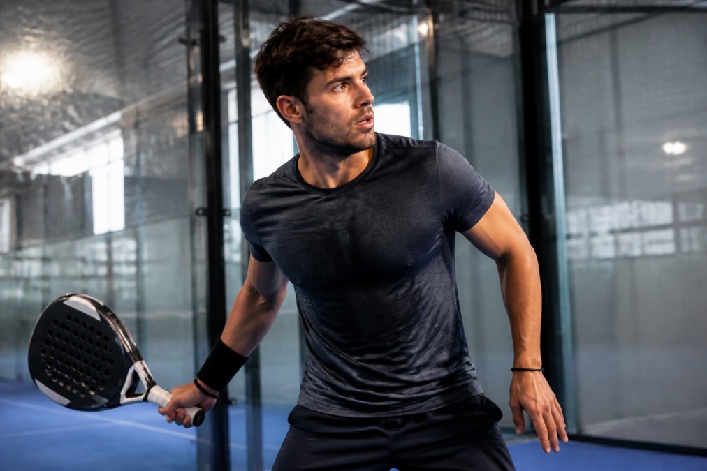 Professional padel player mid-match in modern enclosed court wearing technical moisture-wicking apparel, sweat visible on the fabric as they move dynamically with intense focus