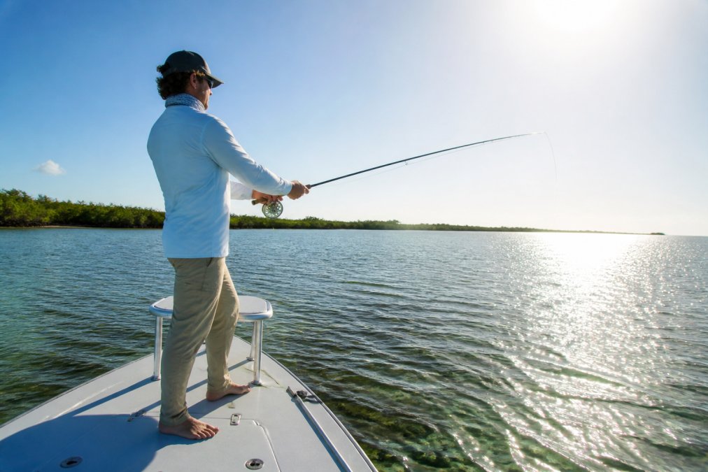 An angler in a long sleeve UPF fishing shirt standing at the bow of a flats boat in bright midday sunlight, casting a fishing rod over open water, with the sun reflecting off the surface around the boat