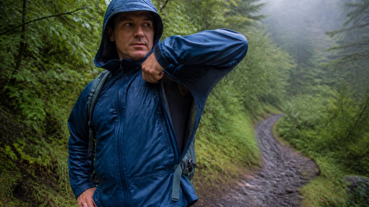 Hiker on a winding trail in humid forest conditions wearing a breathable rain jacket with pit zips partially open, showing ventilation during light rain with mist in the background