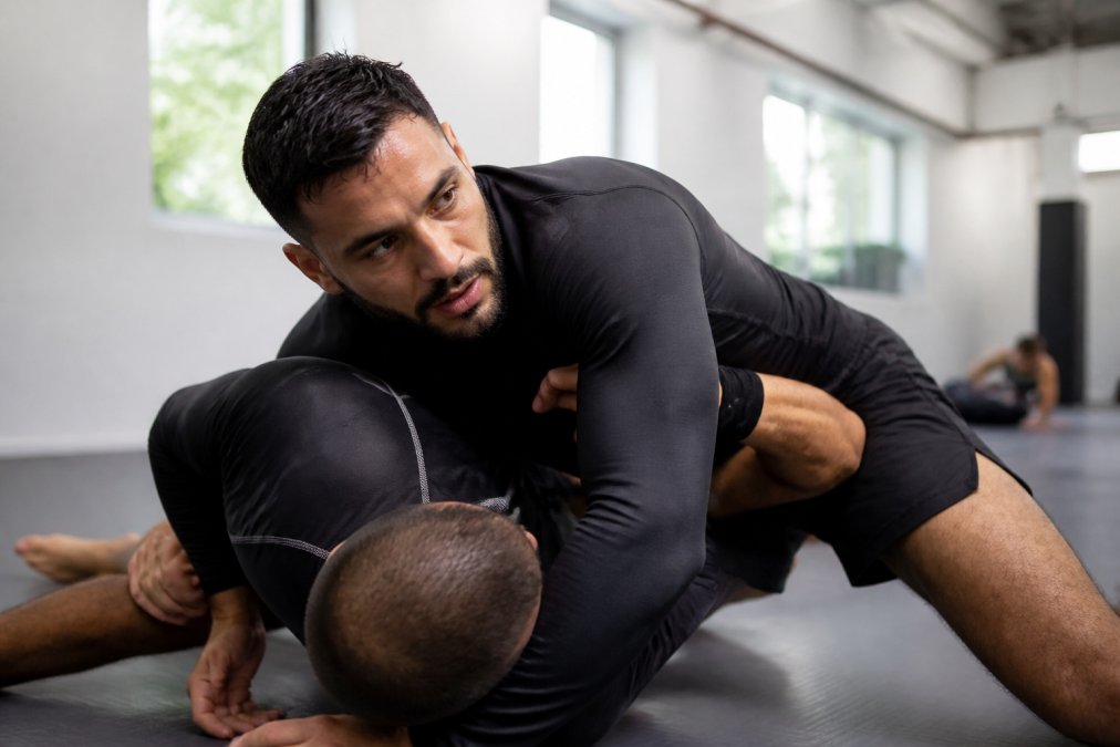 Professional BJJ athlete wearing a breathable rash guard during intense training, grappling with a partner on the mat with natural gym lighting showing the fit and fabric texture in action