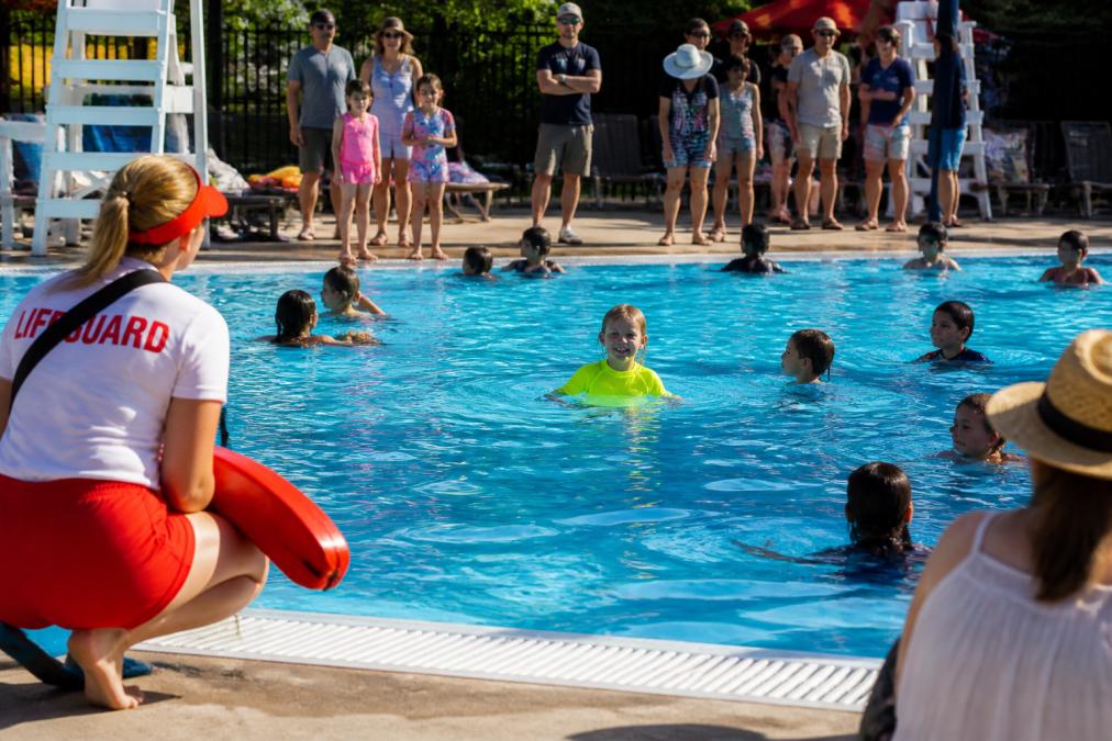 Young child wearing bright neon yellow swimwear in a crowded public pool, clearly visible against blue water while other children in darker swimsuits blend into the background, with lifeguard and parents able to spot the child instantly