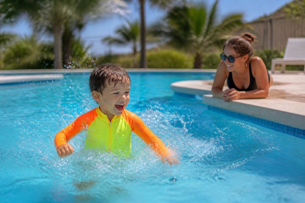 A happy child wearing a bright neon yellow and orange high-visibility swimsuit splashing in a sunny outdoor pool, with a parent watching closely from the edge