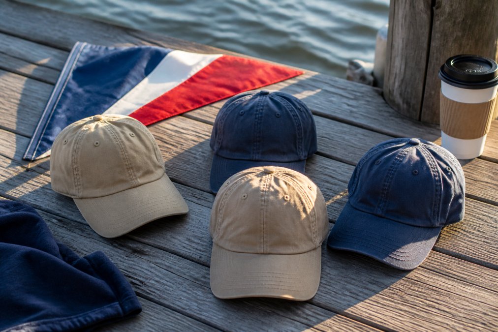 A flat lay of four distressed dad hats in khaki and navy on a weathered wooden dock, with a red, white, and blue pennant and a coffee cup nearby, morning light on the waterfront