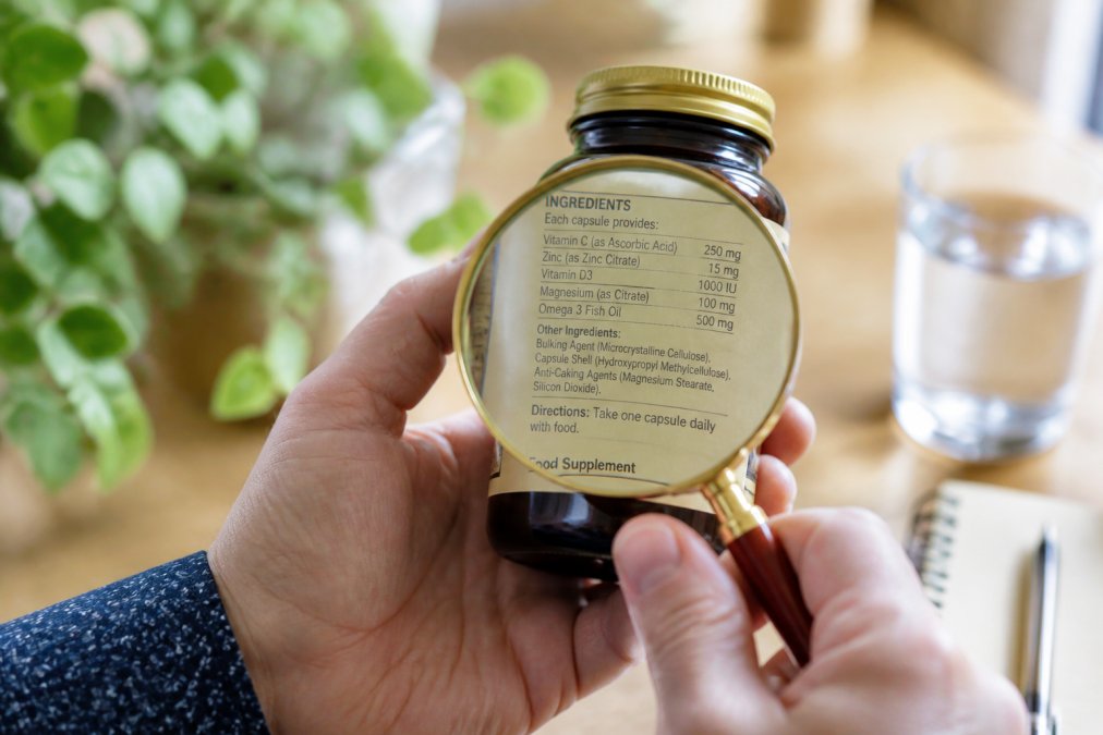 Close-up of hands holding a supplement bottle, examining the ingredient label with a magnifying glass in natural light, showing clearly visible ingredient list and dosage information
