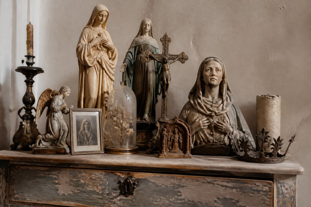 Close-up van antieke religieuze beelden en devotionalia op een vintage kast, met warm licht dat de patina en ouderdomssporen benadrukt