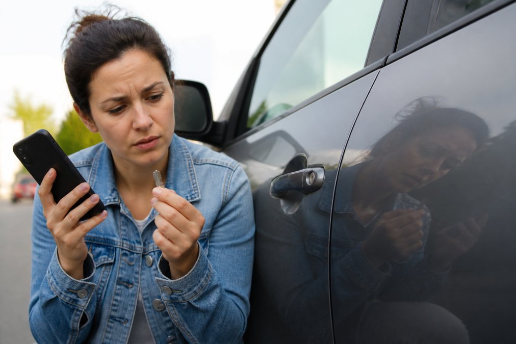A person holding the broken half of a key next to a car door lock, looking concerned, with a smartphone in the other hand ready to call for help