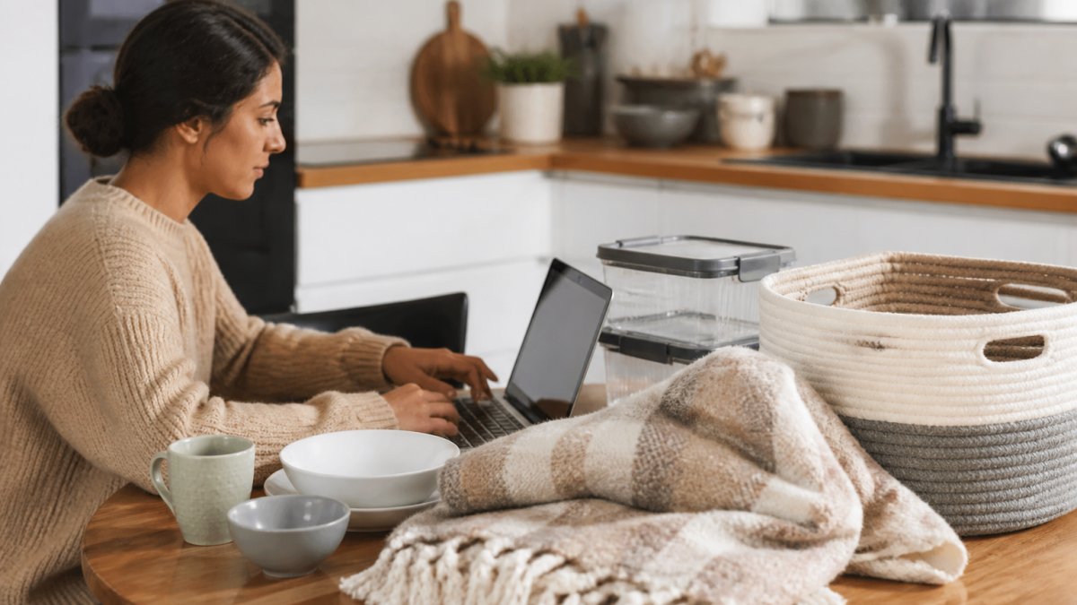 A person browsing home goods on a laptop at a kitchen table, with a cozy throw blanket and storage bins visible nearby, conveying online shopping for home products