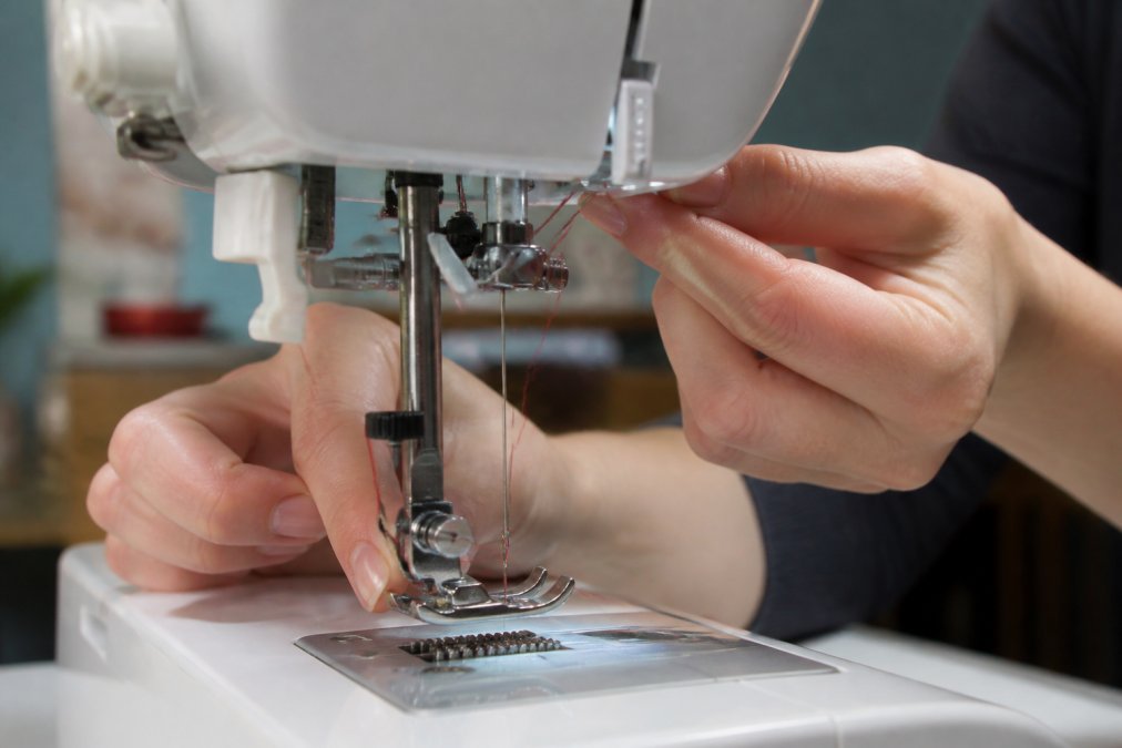Close-up of hands threading a sewing machine needle with the presser foot raised, showing the thread path through the tension discs and needle eye with natural workshop lighting
