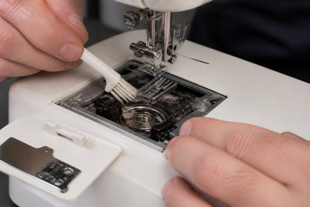 Hands using a small soft-bristled brush to clean the bobbin area of a sewing machine, with lint being removed from the feed dogs and bobbin case, shown in natural workshop lighting