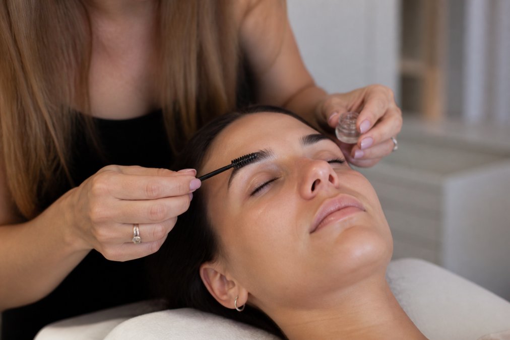 Professional applying brow lamination solution with a spoolie brush on client's eyebrow, showing precise technique and product placement with natural lighting