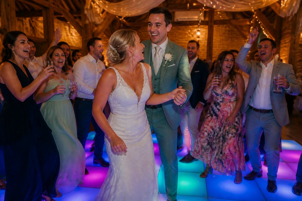 Een lachend bruidspaar danst met hun gasten op een kleurrijk verlichte dansvloer tijdens de receptie, feestelijke ambiance met warme sfeerverlichting
