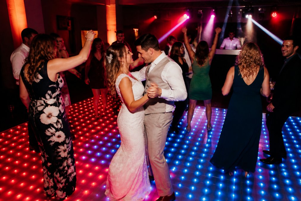 Bruidspaar en gelukkige gasten dansend op een verlichte dansvloer met dynamische rode en blauwe LED-effecten, terwijl de DJ in het achtergrond achter de draaitafel staat met warm licht dat over de menigte speelt
