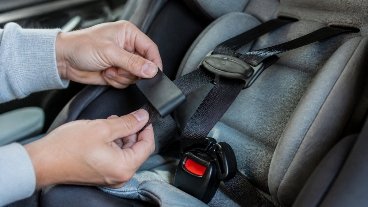 Close-up of a parent's hands sliding a slim silicone sleeve onto a car seat harness strap, with the buckle lying flat and upright in a rear-facing car seat