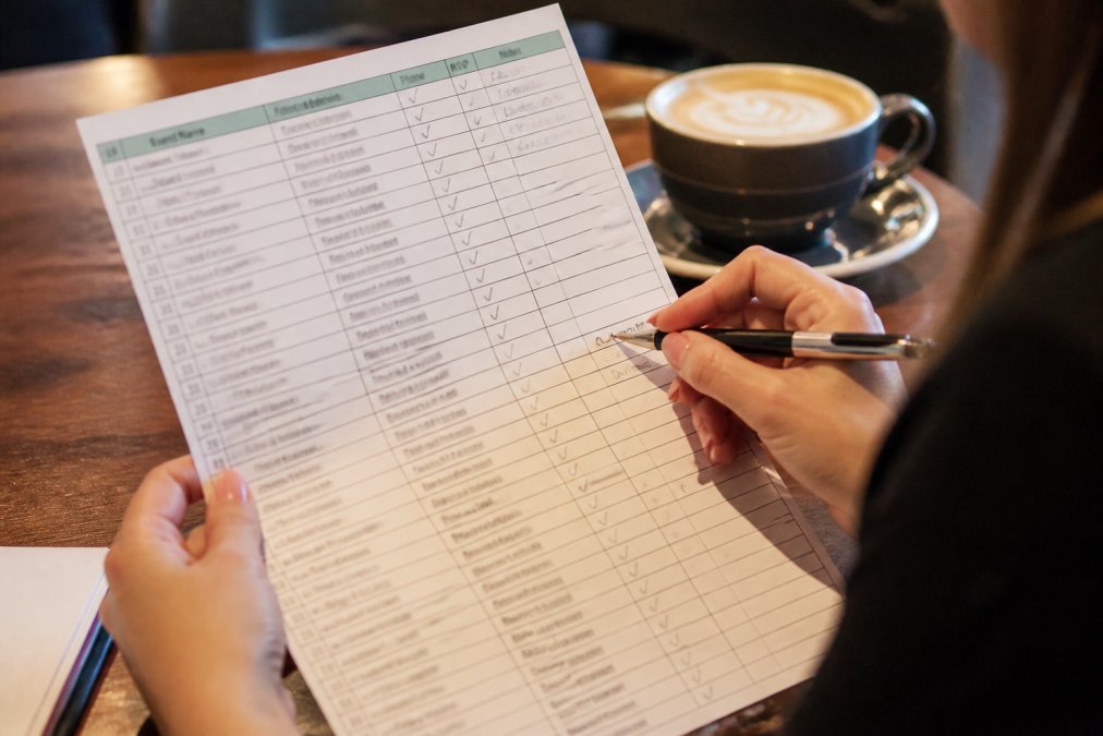 Close-up of hands holding a printed guest list spreadsheet with a pen, coffee cup nearby on a table, showing someone marking off RSVPs