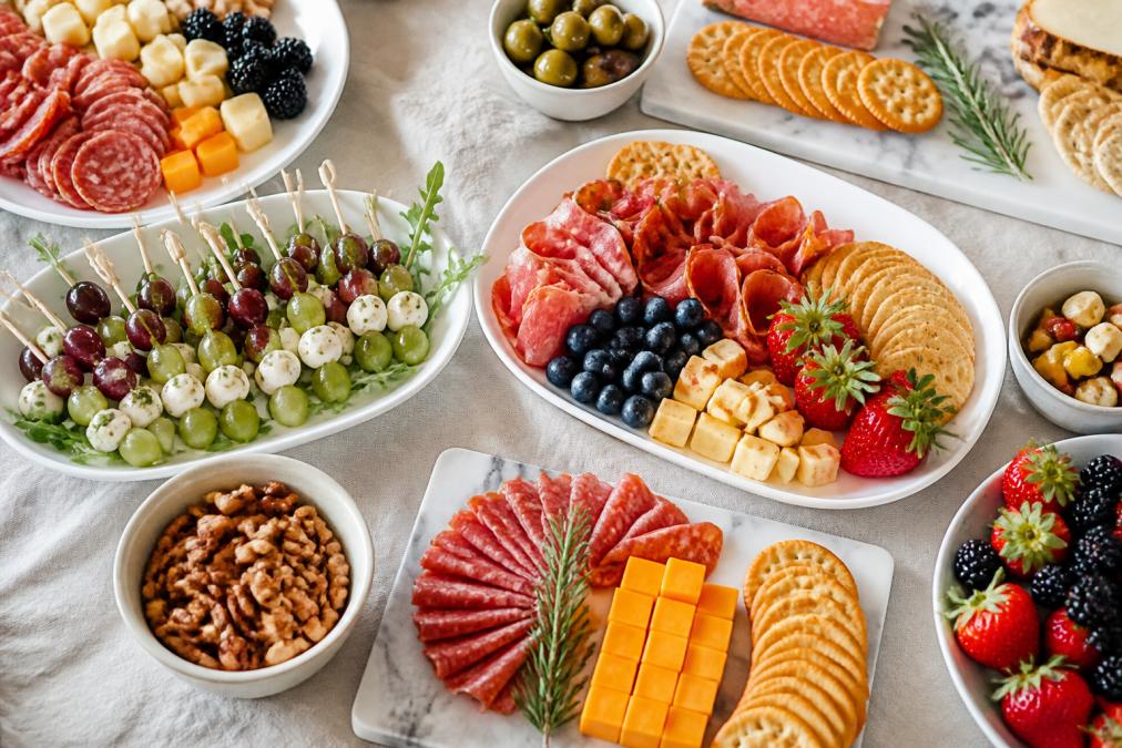 Overhead shot of an elegant appetizer spread featuring multiple white and marble platters arranged with colorful charcuterie, fresh berries, sliced cured meats, assorted crackers, cheese cubes, skewers with olives and grapes, and fresh herbs as garnish on a light linen table setting