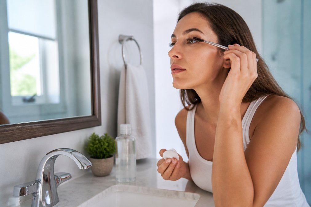 Woman with lifted lashes gently brushing her lashes with a metal spoolie while standing at a bathroom sink, using an oil-free micellar water solution on a cotton pad, soft morning light streaming through the window