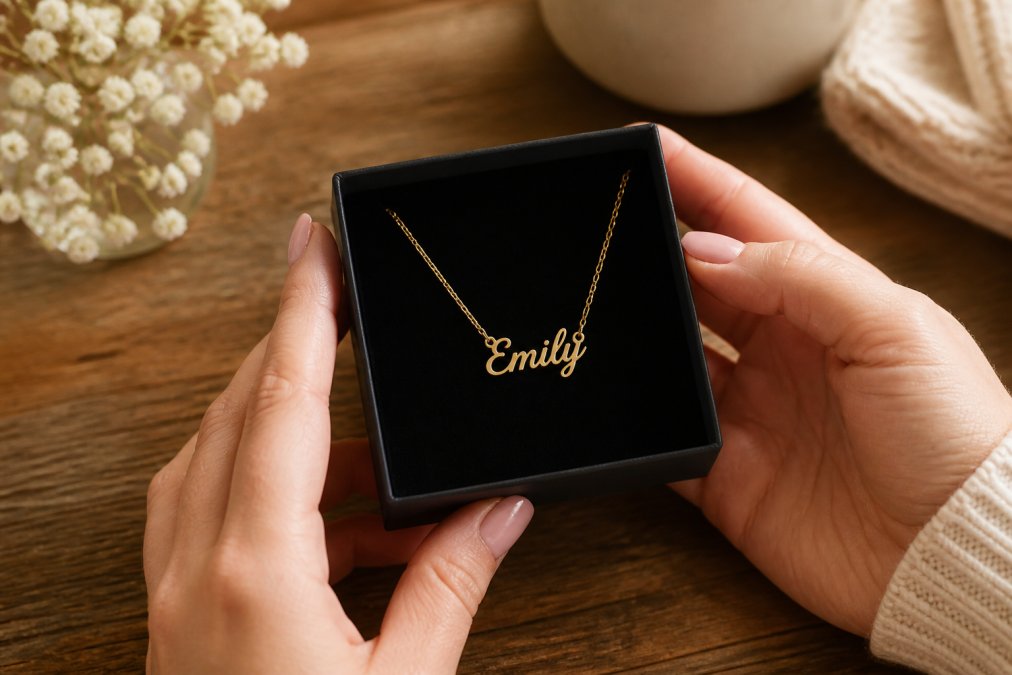 Close-up of a woman's hands holding a small custom name necklace in a soft-touch gift box on a wooden table with warm natural lighting