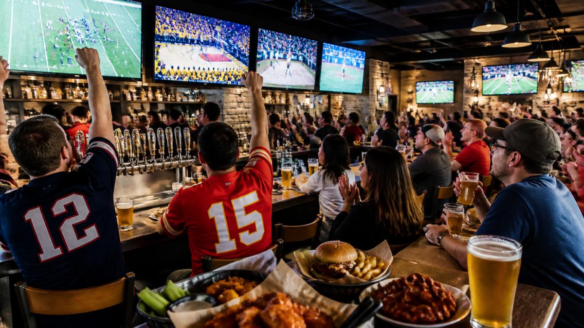 A lively sports bar interior with multiple big-screen TVs showing different games, a packed bar counter with beer taps and baskets of chicken wings, and fans in team jerseys cheering under warm pendant lighting