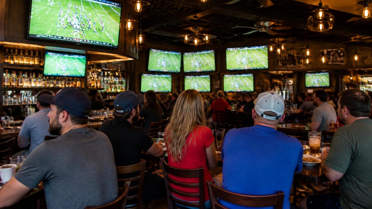 Sports bar interior with multiple large flat-screen televisions mounted on walls at varying heights, patrons seated at high-top tables and bar stools watching a football game, warm amber lighting from pendant fixtures, and casual crowd atmosphere