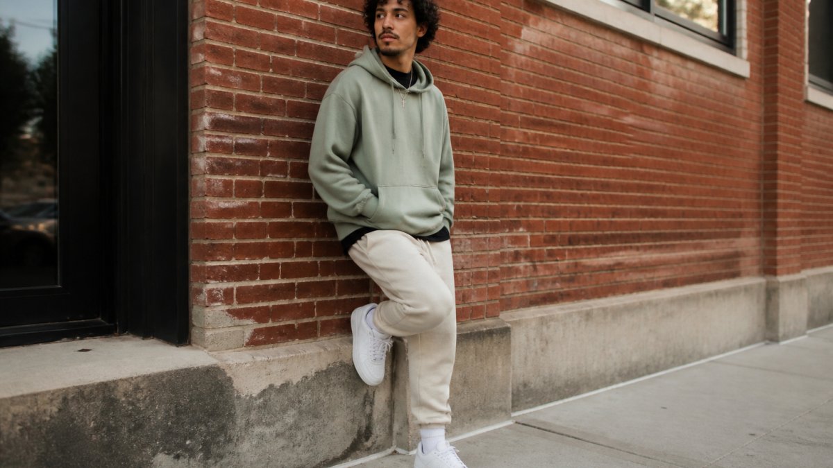 Young adult wearing layered Christian streetwear, black graphic tee visible under an oversized sage green hoodie with cream-colored joggers, posed confidently in an urban setting with brick walls and natural daylight, showing how faith-inspired pieces create a cohesive everyday look
