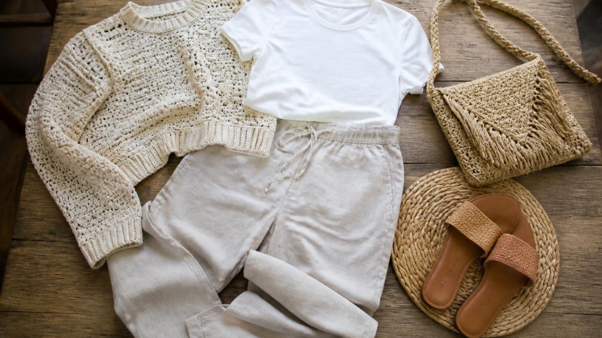 Flat lay of coordinating neutral-toned clothing pieces including a handmade crochet sweater in cream, linen pants in beige, white basic tee, and woven accessories arranged on a wooden surface to show how pieces work together