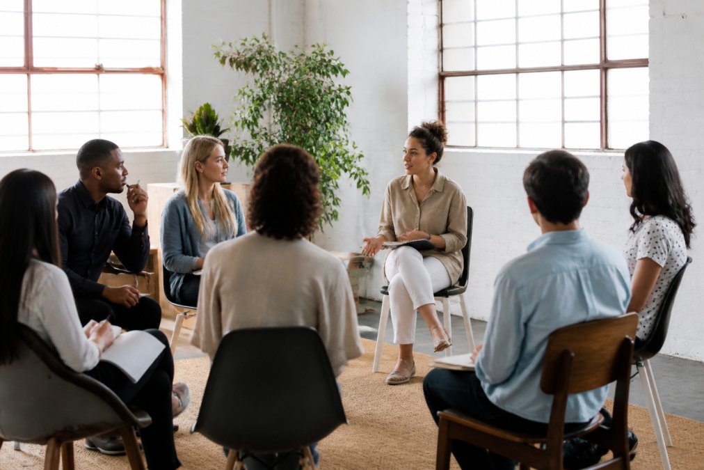 Professional coach facilitating engaged group coaching session with diverse participants in collaborative learning environment, natural light from windows, participants taking notes and engaged in discussion