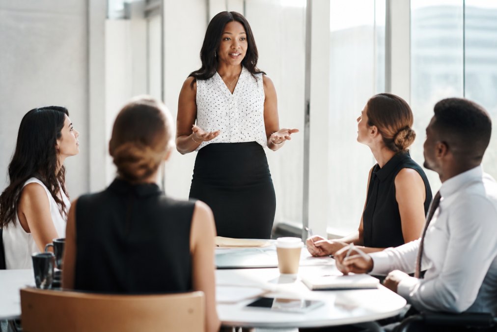 Professional woman speaking confidently during a team meeting in a modern office, colleagues listening attentively with natural light streaming through windows