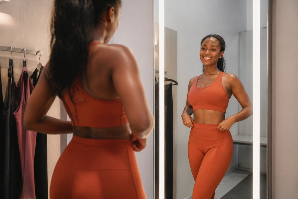 Woman confidently trying on bold-colored activewear in a bright fitting room, smiling at herself in the mirror